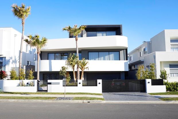 Street-facing view of a contemporary multi-storey residence with crisp white rendered walls, dark window frames, a rounded projecting balcony, broad glazing, a flat roof, and a black slatted front gate. Palm trees and low planting line the frontage, with neighbouring modern homes visible on either side.