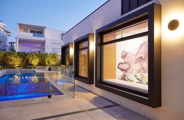 Contemporary low-rise residential exterior at dusk, featuring a tiled terrace beside a blue-lit swimming pool, frameless glass pool fencing, and bold black-framed floor-to-ceiling windows. Warm interior lighting reveals a furnished room and decorative wall art through the glazing, while a neighbouring modern residence and planted screening are visible in the background.