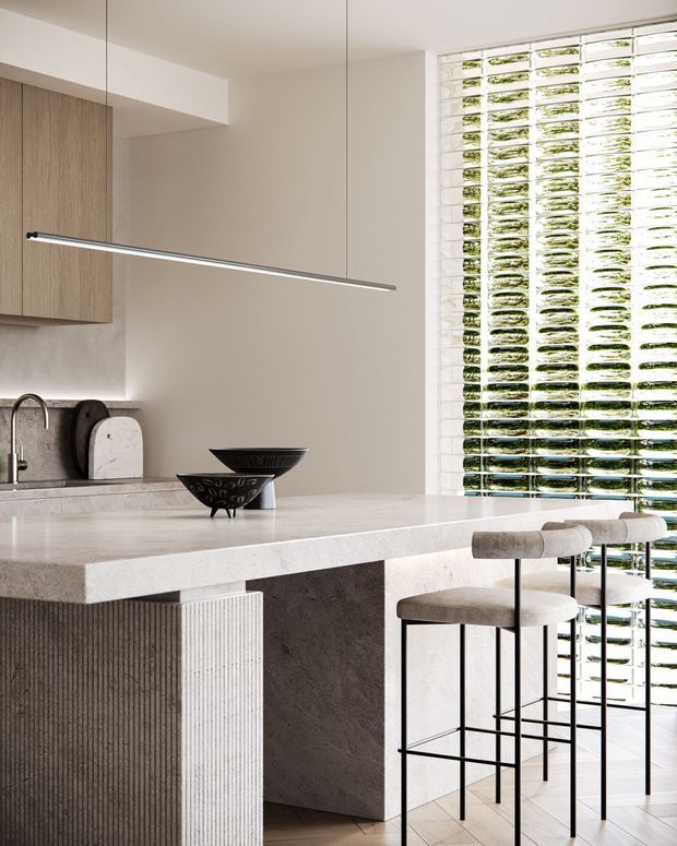 A bright, refined contemporary kitchen centred on a large pale stone island with integrated seating. The island features a fluted vertical detail and a dark textured stone support, while warm wood cabinetry lines the rear wall. A slim linear pendant light hangs above the island, and a full-height glazed brick or textured glass screen brings in filtered natural light and greenery. Neutral materials, sculptural black bowls, and upholstered bar stools create a minimalist, sophisticated interior.