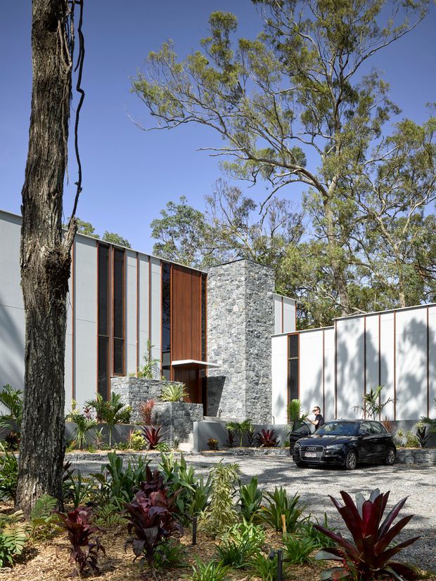 Exterior view of a contemporary multi-level residence set among mature trees and tropical landscaping. The facade combines crisp white and grey panels, tall narrow glazing with dark frames, warm vertical timber cladding, and a prominent rough grey stone-clad entry volume. A gravel driveway and landscaped garden occupy the foreground, with a parked car near the entrance.