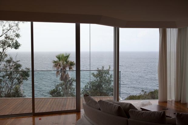 A refined living room overlooking the ocean through expansive floor-to-ceiling glazing. The room features a large curved sectional sofa, warm timber flooring, a timber deck outside, sheer curtains, and a low timber coffee table. Coastal vegetation and open water are visible beyond the glass.