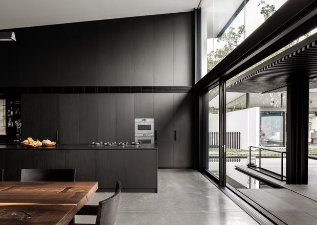 A dramatic modern kitchen and dining area with floor-to-ceiling black-framed glazing opening to an outdoor covered space. The kitchen features dark full-height cabinetry, a long dark island with integrated appliances and cooktop, polished concrete flooring, and a substantial timber dining table with dark chairs. Strong linear geometry, black finishes, and abundant natural light create a refined indoor-outdoor setting.