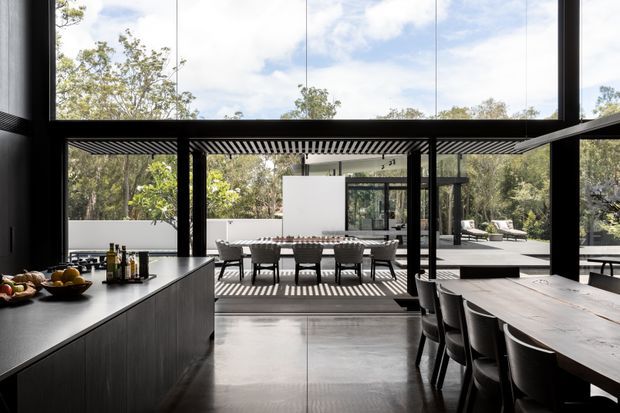 Open-plan contemporary kitchen and dining area with a dark island and integrated cabinetry in the foreground, a large timber dining table to the right, and full-height glazing opening to a covered outdoor dining terrace and landscaped setting. Strong black structural framing, a slatted pergola, polished concrete flooring, and abundant natural light create a seamless indoor-outdoor connection.