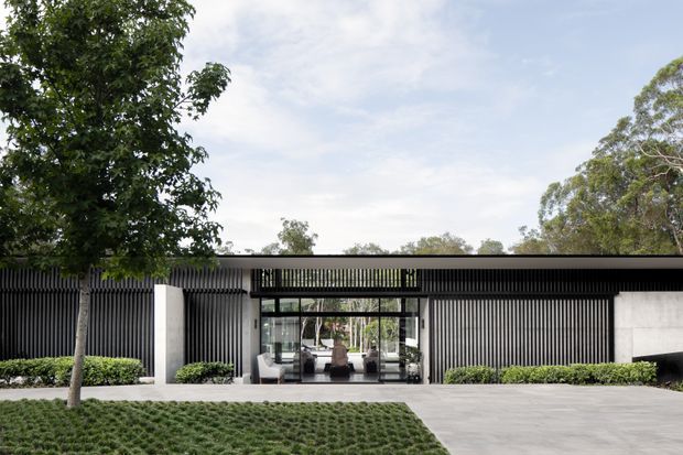 A low, contemporary single-storey building facade set within landscaped grounds. The frontage combines dark vertical screening battens, pale rendered or concrete wall panels, extensive glazed entry doors, and a broad flat roofline. A paved forecourt, clipped hedges, lawn, and mature trees frame the entrance.
