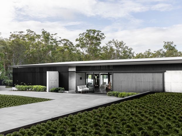 A low, single-storey contemporary building set within a landscaped site. The facade combines dark vertical ribbed cladding, pale concrete or rendered wall panels, extensive glazing and a broad shallow roof. A large paved path leads through clipped groundcover planting to the recessed glazed entry, with dense trees forming the backdrop.
