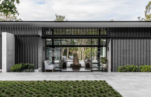 Contemporary single-storey residential facade with a broad flat roof, dark vertical slatted screening, pale rendered or concrete walls, and expansive black-framed glazing. The central glazed opening reveals a calm interior seating area, while a light paved forecourt and neatly planted lawn frame the entrance. The composition is minimalist and strongly horizontal, with a sheltered entry and private landscaped setting.