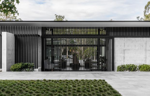 Contemporary single-storey residential facade with a flat roof, dark vertical screening, exposed concrete walls, and a gated entrance. The layered slatted frontage partially reveals a landscaped courtyard and entry beyond, with pale stone paving and low hedging.