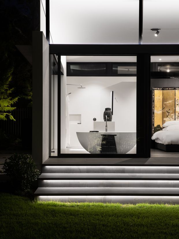 Night-time exterior view of a contemporary home, focused on a fully glazed bathroom with a freestanding bathtub and visible bedroom beyond. Bold black window framing, white interior surfaces, and illuminated exterior steps create a dramatic modern composition.