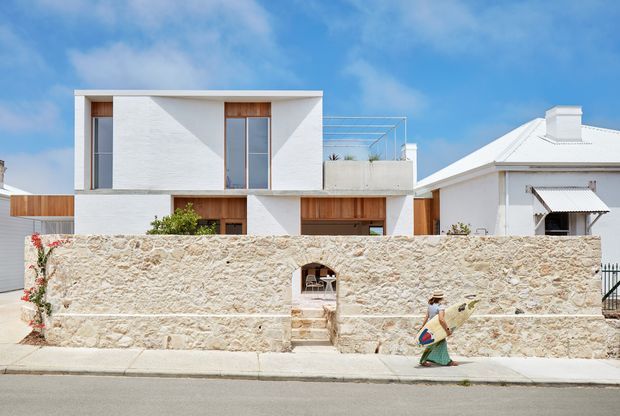 Street-facing view of a contemporary two-storey residence with a crisp white rendered facade, tall narrow windows, warm timber-clad recessed openings, and a rooftop terrace with a slender metal balustrade. A textured pale stone boundary wall with a small pedestrian gate fronts the property. A neighbouring traditional white house with a pitched metal roof is visible to the right, under a bright blue sky.