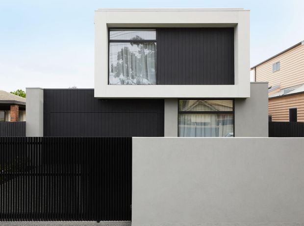 Street-facing view of a contemporary two-storey residence with a minimalist cubic form. The facade combines smooth light-grey rendered walls, dark vertical cladding, large rectangular windows, and a tall black vertical-slat front fence. Adjacent suburban houses are partially visible on either side.
