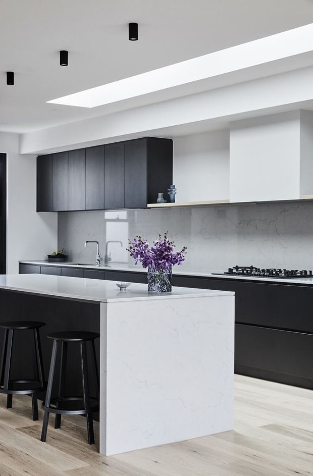 A bright contemporary kitchen with a large white stone island, dark charcoal cabinetry, a matching dark cooking run, pale timber flooring, and a light stone backsplash. Black bar stools sit at the island, while a long linear skylight and recessed black cylindrical downlights illuminate the space.