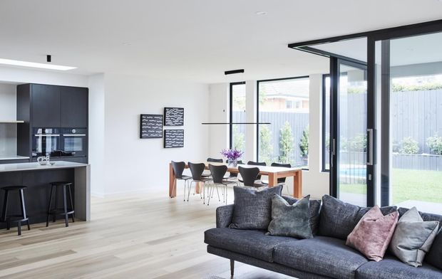 Bright open-plan contemporary living, dining and kitchen interior with pale timber flooring, white walls, dark kitchen cabinetry, a stone island with bar seating, a compact dining setting, and a dark grey sofa in the foreground. Large black-framed glazed doors open toward a landscaped backyard, creating an airy connection to the outdoors.