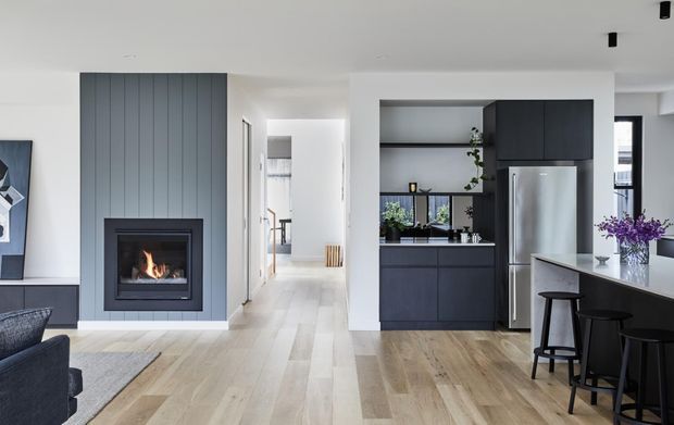 A bright open-plan contemporary interior combining a living area, kitchen and dining or breakfast bar. The room features a dark charcoal vertical-panel fireplace surround with an inset fire, dark flat-panel kitchen cabinetry, stainless-steel appliances, a white island benchtop, pale timber flooring, and large windows providing natural light. A hallway is visible beyond the living space.