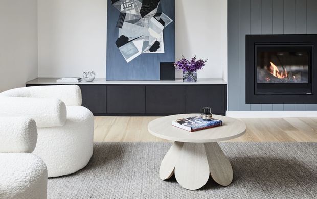 A refined contemporary living room featuring a sculptural cream boucle armchair and matching seating, a pale round coffee table with a petal-like base, a large textured neutral rug, and a dark built-in media console. A modern black-framed fireplace is integrated into grey vertical wall panelling, with abstract artwork and a small floral arrangement adding visual interest.