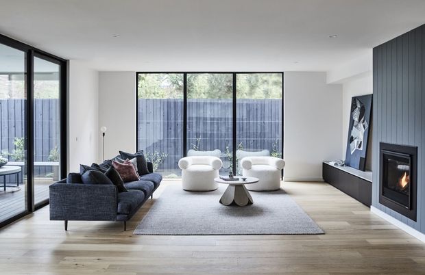 Bright contemporary living room with pale timber flooring, white walls, expansive black-framed glazing, a dark sectional sofa, two rounded white lounge chairs, a low sculptural coffee table, and a built-in dark fireplace surround with artwork. The room opens visually to a fenced outdoor area.