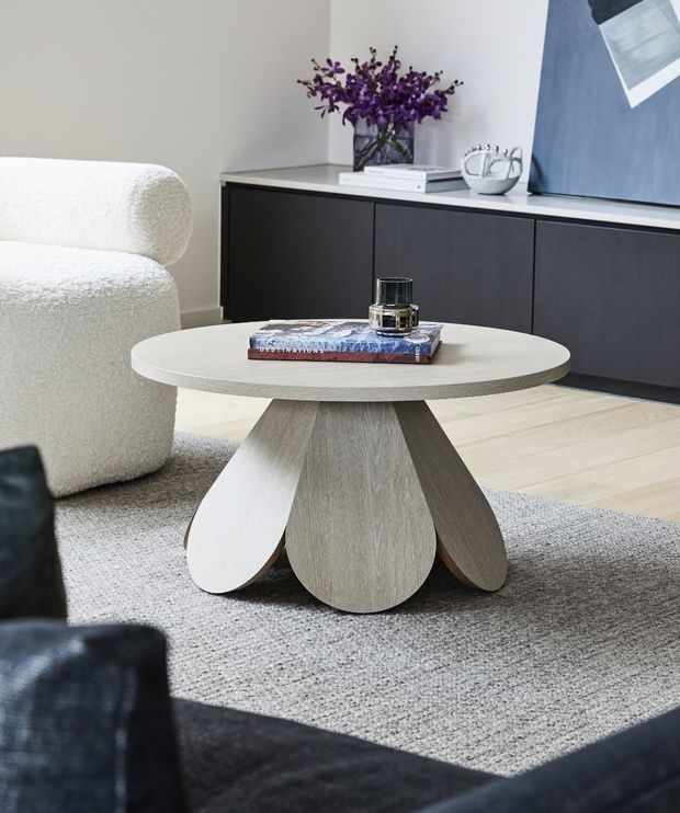 A refined contemporary living room vignette centered on a round light-wood coffee table with a sculptural, petal-like pedestal base. The table sits on a textured grey rug beside a boucle upholstered armchair, with dark built-in cabinetry, pale timber flooring, abstract artwork, books, glassware and purple flowers in the background.