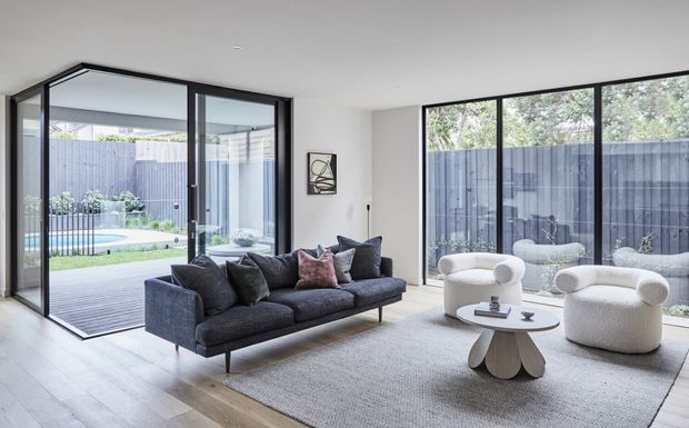 Bright contemporary living room with floor-to-ceiling black-framed glazing and sliding doors opening to a timber deck, landscaped yard, and pool. The room features a charcoal sectional sofa, two rounded cream lounge chairs, a sculptural white coffee table, and a large neutral rug.
