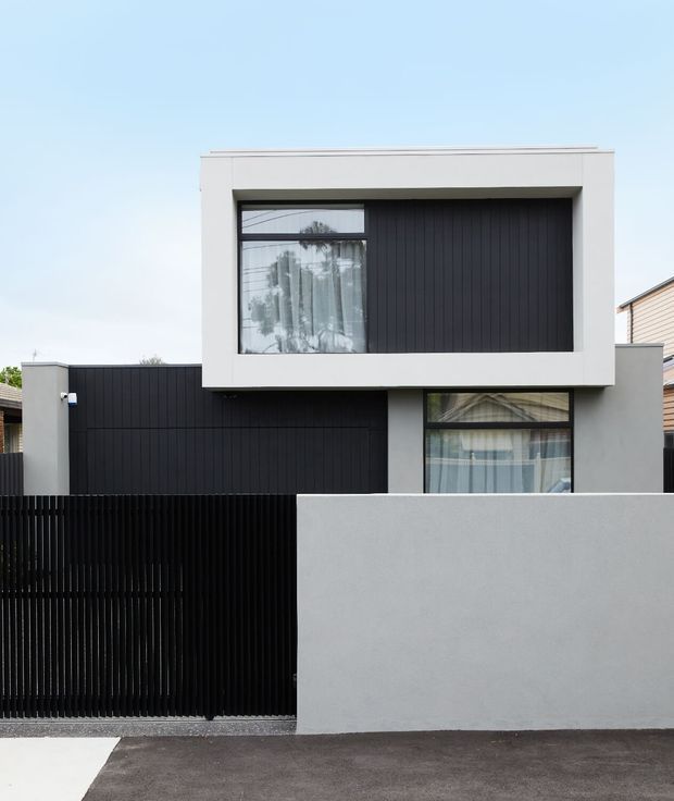 Front exterior view of a contemporary two-storey urban residence with a strong geometric composition. The facade combines smooth white and light-grey rendered wall planes with dark vertical cladding, a projecting upper-level box, large glazed openings, and a tall black slatted front fence and gate. Neighbouring homes are visible at the edges.