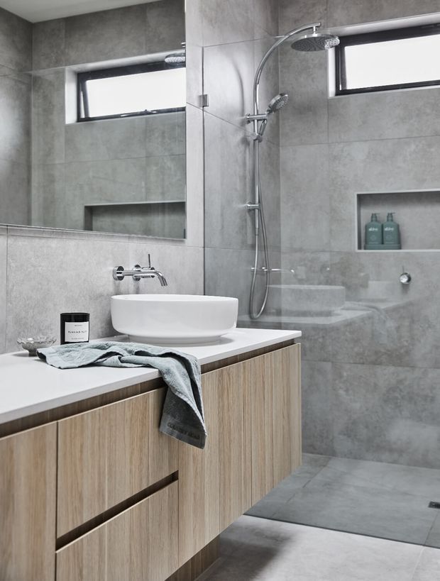 Contemporary grey tiled bathroom with a large frameless mirror, floating light timber vanity cabinetry, white vessel basin, and chrome wall-mounted tapware. A walk-in shower with dual shower heads, recessed wall niche, and high horizontal windows occupies the background. The restrained palette and clean lines create a refined, modern atmosphere.