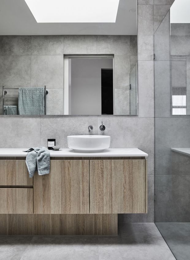 Contemporary bathroom vanity featuring a floating timber-look cabinet with vertical grain, white vessel basin, pale benchtop, large grey concrete-look wall and floor tiles, and a frameless glass shower enclosure. A wide mirror, skylight-like ceiling opening, heated towel rail and muted teal towels complete the clean, refined scheme.