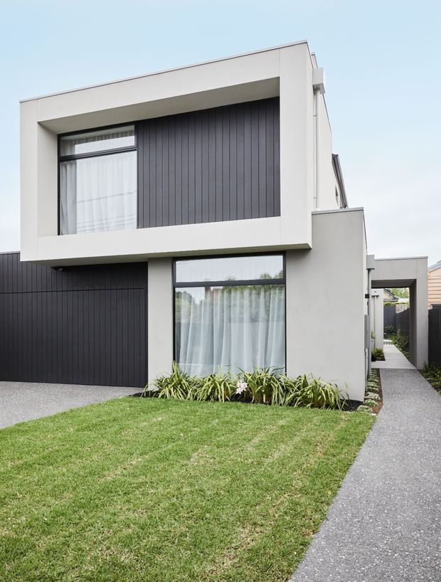 A contemporary two-storey residential facade with a crisp white rendered frame, dark vertical cladding, large glazed windows, and a projecting upper-level box. A neat lawn, planted garden bed, driveway, and narrow paved side path occupy the foreground.