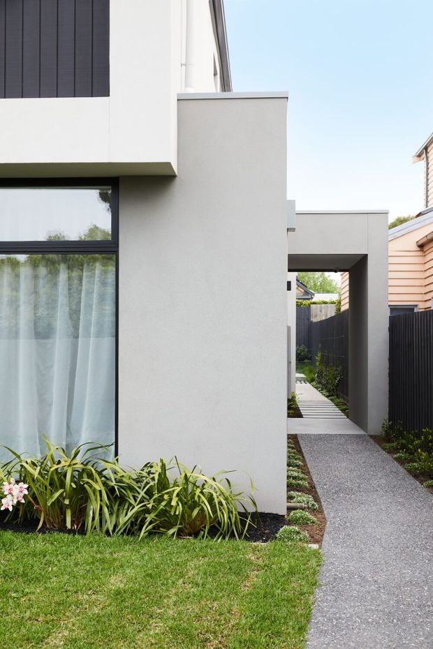 A contemporary residential exterior with clean white rendered walls, dark-framed glazing, and strong rectilinear forms. A narrow grey gravel path runs alongside the building and passes beneath a projecting covered walkway or portal. Black fencing, simple garden planting, and a neighbouring weatherboard house are visible.