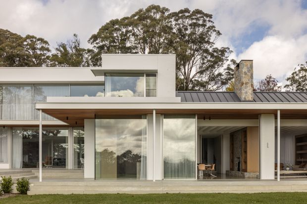 A contemporary two-storey residential facade with crisp white rendered volumes, extensive floor-to-ceiling glazing, slender white columns, and a broad timber-lined covered terrace. The low-pitched dark metal roof includes a prominent natural-stone chimney. The house opens onto a landscaped lawn, with mature trees visible behind it.