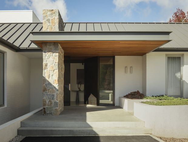 Single-storey residential entry facade with a recessed front door beneath a broad timber-lined soffit. The entrance is framed by a substantial irregular natural-stone pillar, pale rendered walls, dark standing-seam metal roofing, concrete steps and a raised white planter. Glazing and a partially visible interior occur behind the entry, while the overall composition is clean and contemporary with strong horizontal lines.