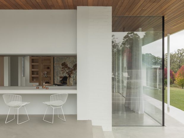 A refined modern interior showing a minimalist kitchen or bar counter with two wire-frame stools, a white tiled structural column, polished concrete flooring, and expansive floor-to-ceiling glazing opening toward a landscaped garden. The ceiling is clad in warm timber boards, creating contrast with the predominantly white and grey palette. Reflections reveal additional interior furnishings and a dining or living area.