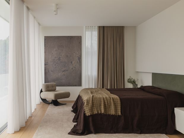 A refined contemporary bedroom with floor-to-ceiling sheer curtains along one wall, layered taupe and brown drapery, a low platform bed dressed in deep brown linen, and a pale upholstered lounge chair. Light timber flooring and a large neutral rug create a warm, muted base, while a textured abstract wall artwork and soft concealed-looking bedside joinery add understated character.
