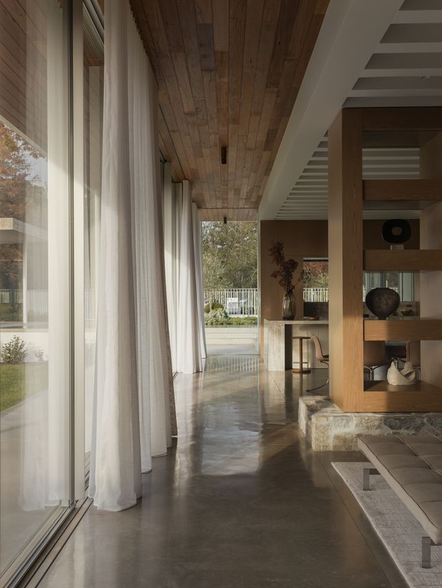 A refined open-plan interior hallway and living area with floor-to-ceiling glazing, sheer white curtains, a polished concrete floor, and a warm timber-clad ceiling. A built-in timber shelving partition frames the living space, with a stone-clad raised hearth or platform and neutral furnishings visible at right. The space opens toward an outdoor terrace and garden, creating a bright, calm indoor-outdoor connection.