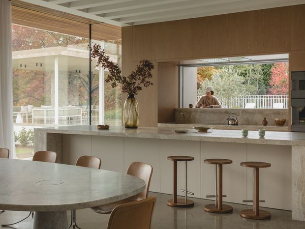 A refined open-plan kitchen and dining area with a long pale stone island, integrated light cabinetry, timber wall panelling, and a large internal serving or viewing opening. Three timber-topped stools line the island, while a rounded stone dining table and curved upholstered chairs occupy the foreground. Floor-to-ceiling glazing connects the room to a leafy outdoor setting, and a white exposed-beam ceiling adds architectural detail. The palette is warm, muted, and natural.