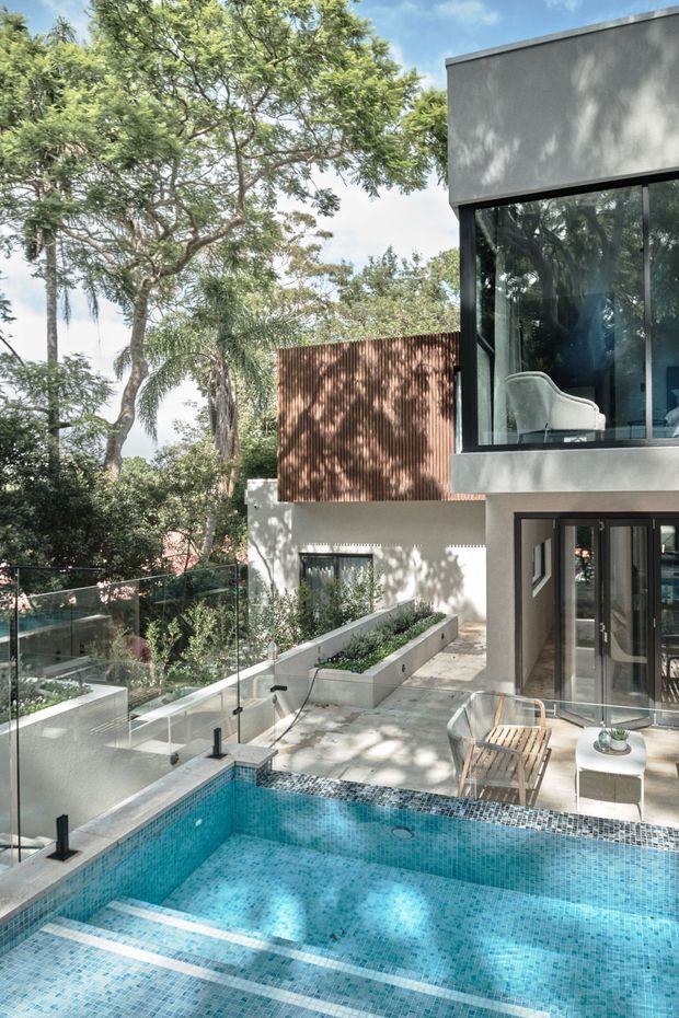 A contemporary multi-level residential exterior with a tiled swimming pool in the foreground, glass balustrades, landscaped raised planters, and large dark-framed windows. The building combines smooth rendered walls with a warm corrugated metal-clad section, surrounded by mature trees and greenery.