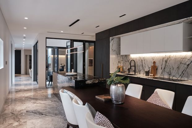 Contemporary open-plan kitchen and dining area with a long dark dining table, white upholstered chairs, black cabinetry, a dramatic white marble backsplash and benchtop, and a central stone island. Polished marble flooring, integrated lighting, glass partitions, and a dark timber-toned feature wall create a refined, high-contrast interior.