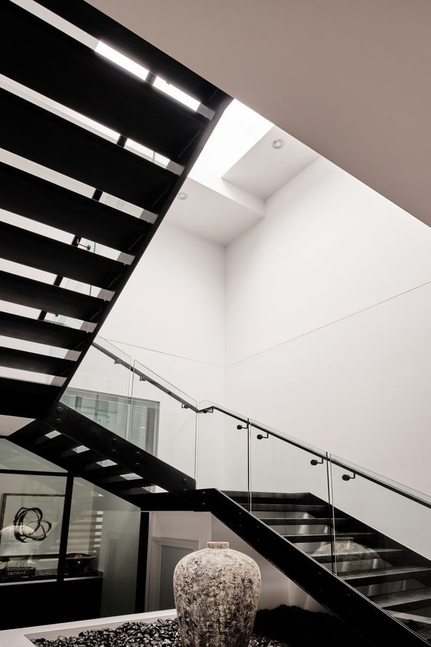 Interior view of a dramatic modern stairway with black open-riser steel treads and a black structural frame, paired with clear glass balustrades and minimal metal fittings. The stair rises through a bright white double-height space with recessed downlights and clerestory-like glazing. A sculptural stone vessel sits in a dark decorative pebble feature at the base, with a glimpse of adjacent glazed interior space below.