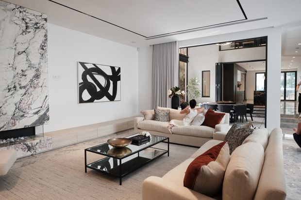 A bright, spacious contemporary living room with two large cream sofas, a black glass coffee table, pale wood built-in media cabinetry, and a dramatic marble-clad fireplace wall. A large black-framed opening and glazing connect visually to an adjacent dining and circulation area. The room features recessed linear ceiling detailing, sheer curtains, neutral upholstery, and rust-red accent cushions.