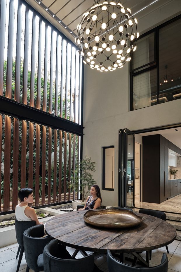 A sophisticated double-height dining area with a large round rustic timber dining table and black upholstered dining chairs. Floor-to-ceiling glazing is screened by tall vertical timber slats, creating a strong connection to greenery outside. A dramatic spherical pendant light hangs overhead, while an open glazed doorway leads toward a contemporary kitchen. The space combines warm timber, dark metal framing, neutral walls, and abundant natural light.