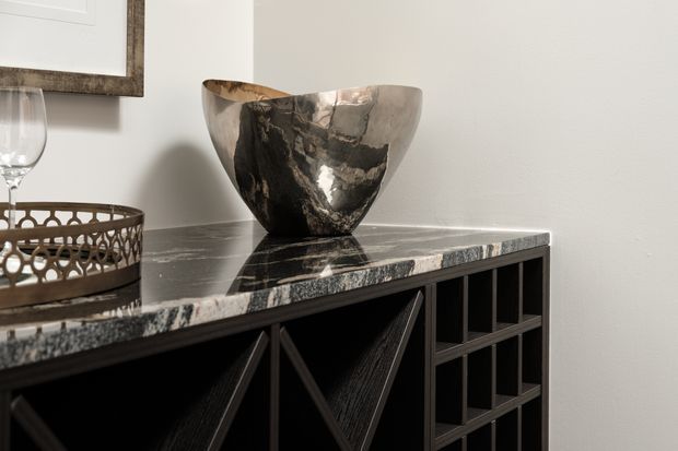 Close-up interior detail of a dark, geometric built-in sideboard or cabinet with open wine-storage cubbies and angular textured doors. The cabinet has a polished black-and-white veined stone benchtop, topped with a sculptural metallic vessel and decorative tray. A neutral light wall and framed artwork form the background.