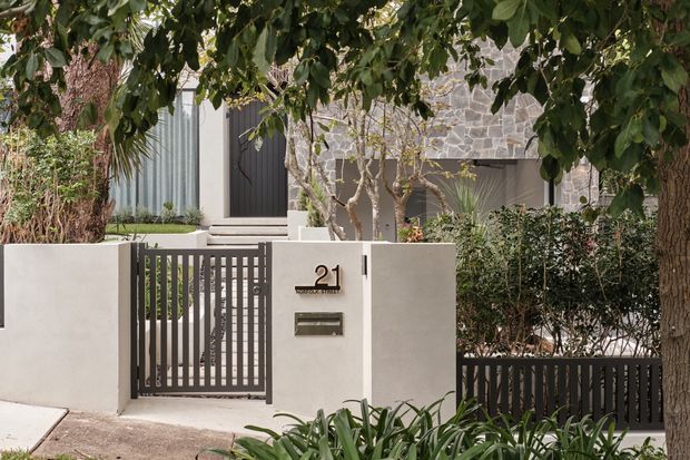 Street-facing view of a contemporary residence entrance, featuring a rendered masonry boundary wall with integrated metal slat pedestrian gate, house number and letterbox. Behind it is a dark entry door, pale stone-clad facade, landscaping, and mature street trees.