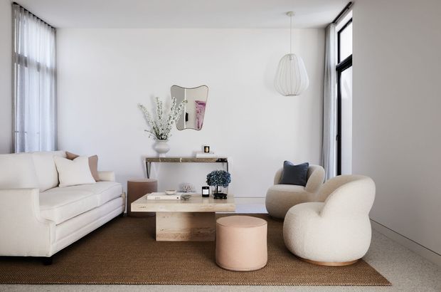 Bright, refined living room with white walls, full-height windows, a cream sofa, sculptural rounded lounge chairs, a pale timber coffee table, and a neutral woven rug. Minimal decor and soft natural light create a calm contemporary interior.