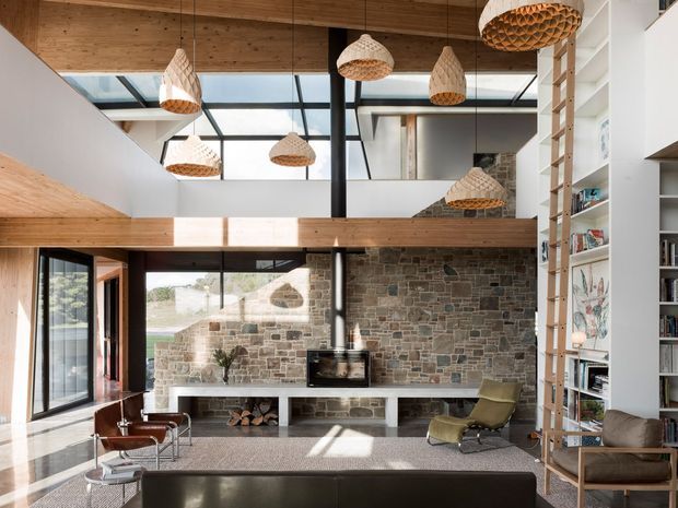 A spacious double-height living room with a dramatic glazed roof and clerestory windows, exposed timber beams, and a substantial natural stone feature wall surrounding a wood-burning stove. The room includes a large dark sofa, leather lounge chairs, built-in shelving, a tall timber ladder, and multiple woven pendant lights. Strong daylight, warm timber, and textured stone create a refined yet natural contemporary interior.