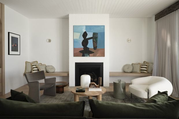 A refined living room viewed from behind a dark green sofa, featuring a central black fireplace set into a white chimney breast, built-in bench seating with striped cushions, sculptural lounge chairs, a curved cream armchair, a timber coffee table, floor-to-ceiling curtains, and a white timber-lined ceiling. Abstract artwork and neutral furnishings create a sophisticated, calm interior.