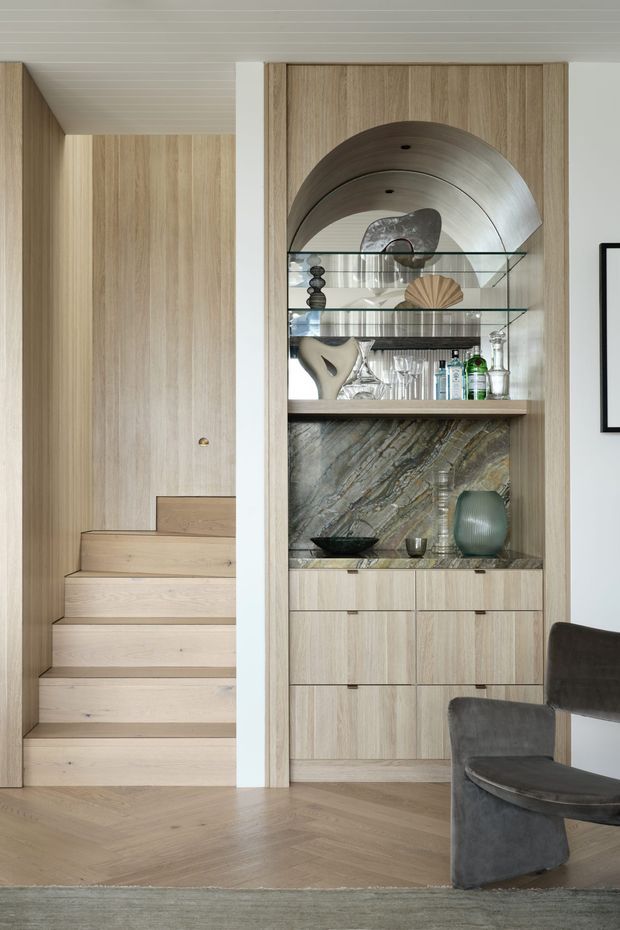 Refined contemporary interior featuring a light timber staircase and matching built-in display cabinetry. The cabinet has an arched metallic-lined niche, glass shelving, a dark veined stone feature backing, and lower drawers. A sculptural dark upholstered chair sits in the foreground, with herringbone timber flooring and a pale horizontally paneled ceiling.