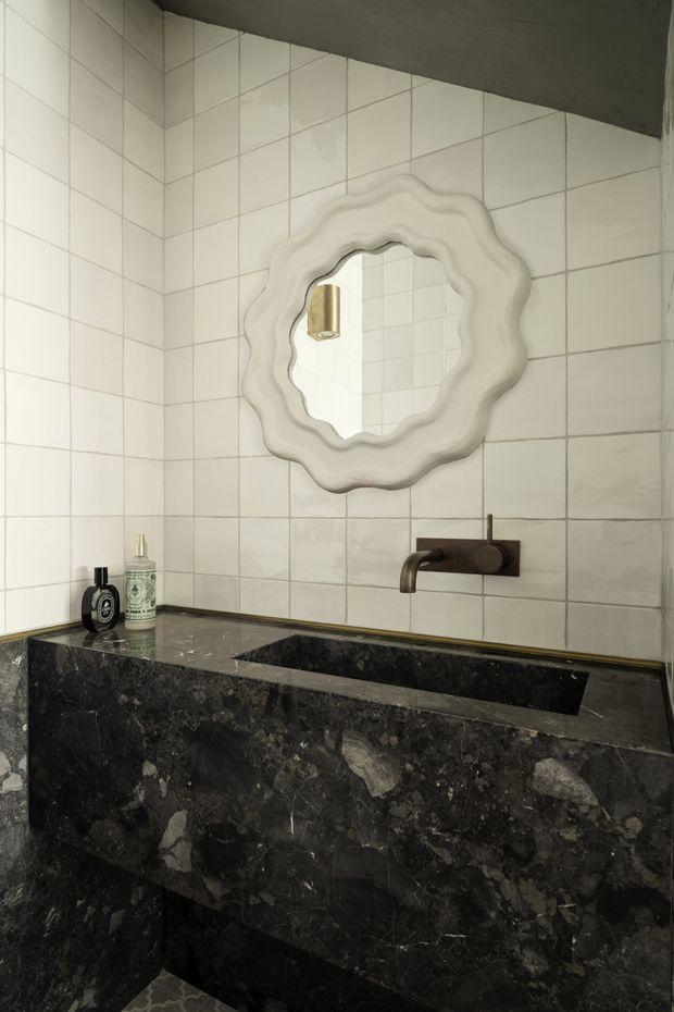 A compact bathroom vanity with a dramatic dark natural-stone basin and matching wall-mounted benchtop, set against off-white square tiled walls. A sculptural scalloped white-framed mirror and aged bronze tapware add a decorative, refined character.