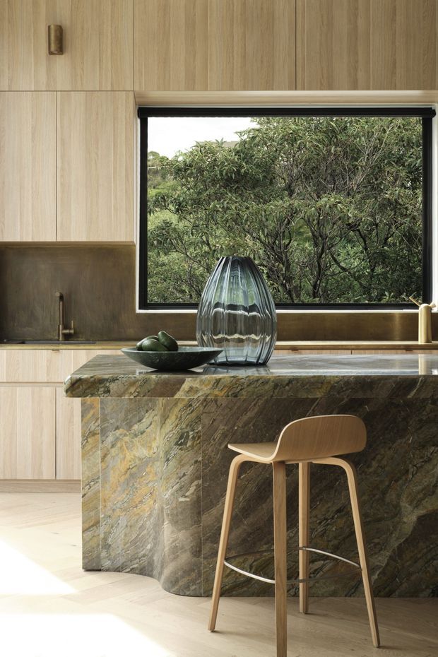 A refined contemporary kitchen featuring light timber cabinetry, a large sculptural natural-stone island with a dramatic veined surface, and a black-framed picture window overlooking dense greenery. A timber bar stool, dark bowl, and ribbed glass vase add warm, organic accents.