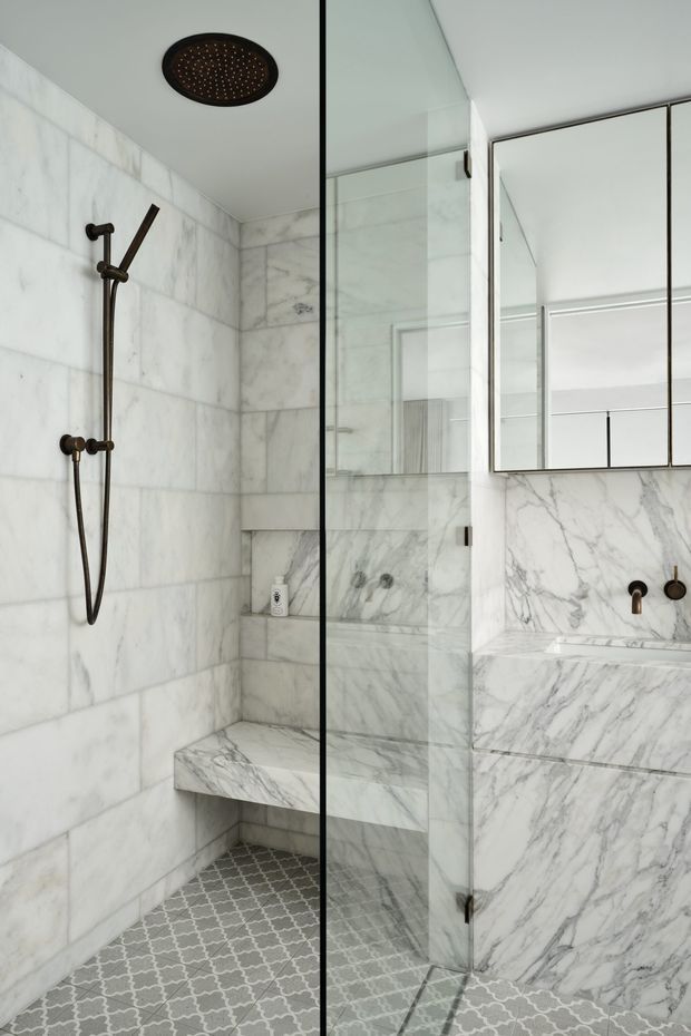 A refined contemporary bathroom featuring a walk-in glass shower with a ceiling-mounted rain shower and wall-mounted hand shower, marble wall cladding and built-in shower bench, patterned grey-and-white floor tiles, and a marble vanity with wall-mounted bronze fixtures and mirrored cabinetry.