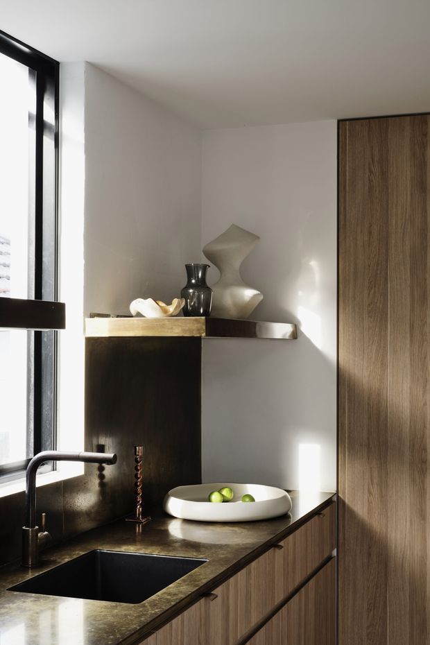 A refined contemporary kitchen corner with dark stone benchtops, integrated cabinetry in warm wood tones, a deep black undermount sink, and a dark metal faucet. A slim floating shelf displays sculptural ceramic objects, while strong daylight enters through a tall black-framed window, creating dramatic light and shadow.