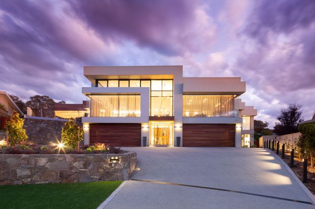 Twilight exterior photograph of a large, contemporary multi-level residence with strong horizontal massing, flat roof planes, extensive floor-to-ceiling glazing, warm interior lighting, and two wide timber-toned garage doors. The home is approached by a broad concrete driveway and framed by stone retaining walls, landscaped planting, and dark fencing.