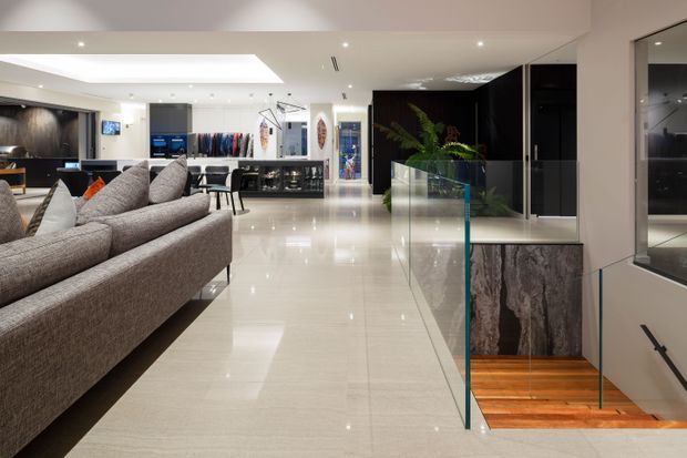 A bright, spacious contemporary open-plan interior featuring a large grey sectional sofa, glossy pale tiled flooring, recessed ceiling lighting, and extensive built-in display and storage joinery. A glass balustrade encloses a stairwell descending to a lower level, with timber stair treads and a dark textured feature wall. The rear area appears to function as a retail or showroom-style space with clothing displays and counters.