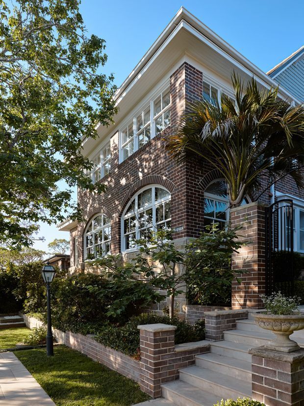 Exterior architectural photograph of a substantial multi-storey brick house, viewed from the landscaped front garden and stair approach. The facade features dark red-brown brickwork, large white-framed arched and rectangular windows, decorative white eaves and trim, a pitched gable roof, raised planting beds, and a formal entry stair. Mature trees and hedges surround the property.